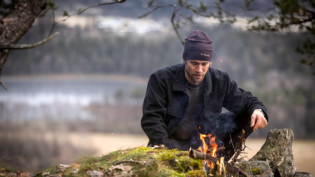 Ein Mann mit Haube und Jacke von Aclima macht ein Lagerfeuer in der Natur.