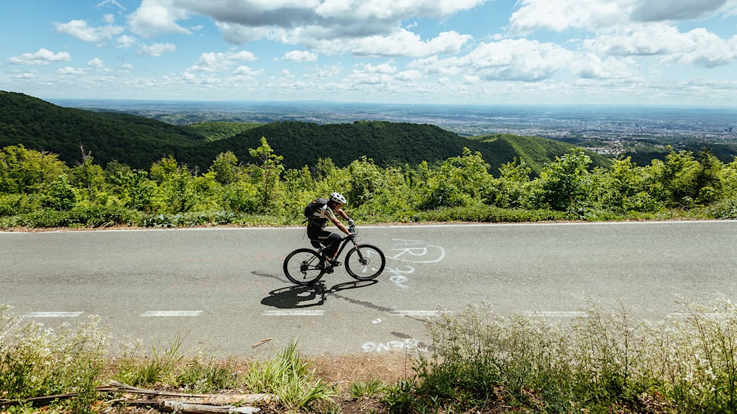 Ein Fahrradfahrer auf der Strecke umgeben von Bergen, Wald und Sonnenschein.