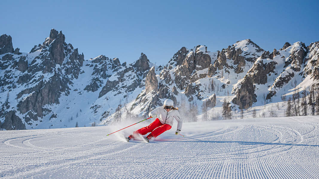 eine skifahrerin fährt eine piste hinunter, der himmel ist strahlend blau und man sieht schneebedeckte bergspitzen im hintergrund