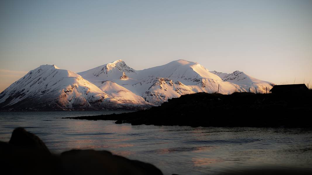 Schneebedeckte Berge der Lyngenalpen, beleuchtet vom warmen Licht der Mitternachtssonne, mit einem ruhigen Fjord im Vordergrund.