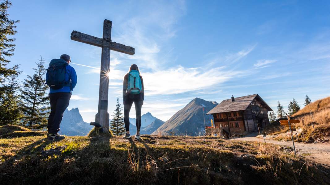 Zwei Wanderer stehen neben einem Gipfelkreuz daneben eine Hütte.