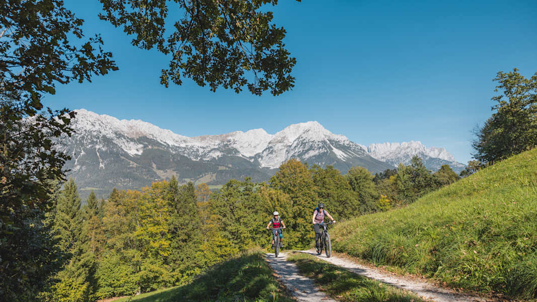 Ein Mann und eine Frau radeln auf einem Forstweg bergauf. Im Hintergrund stehen schneebedeckte Berge.