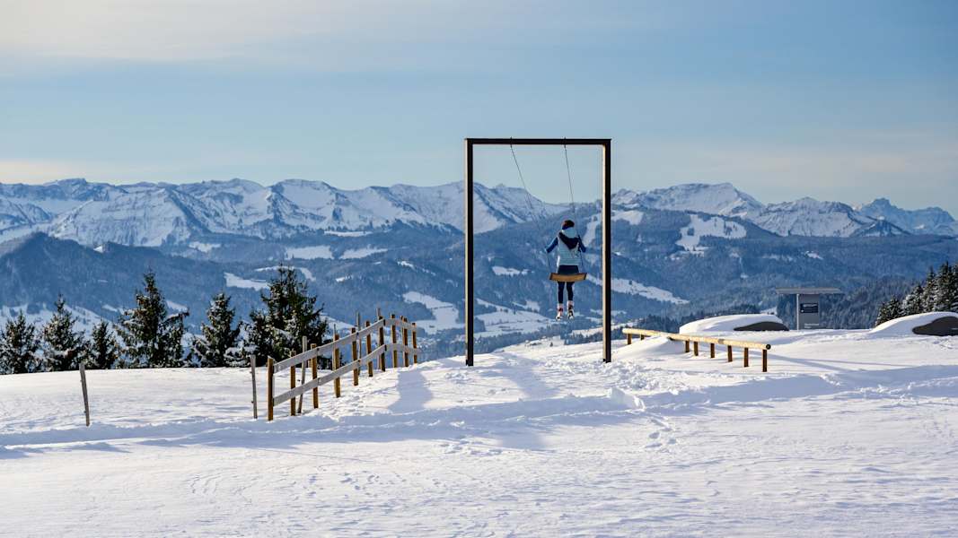 Ein Mädchen schaukelt auf einer Großschaukel vor verschneiter Landschaft. Im Hintergrund die Allgäuer Alpen.