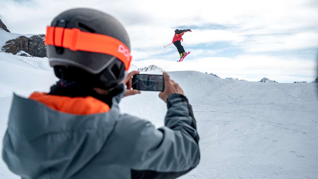 Ein Skifahrer macht einen Stunt über einen kleinen Schneehügel, während ihn ein anderer Skifahrer dabei filmt.