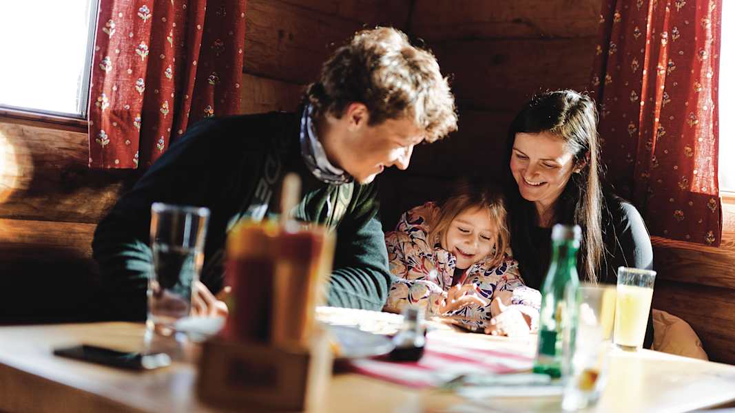 Eine dreiköpfige Familie sitzt in einer Skihütte und sucht auf der Speisekarte das passende Essen aus.