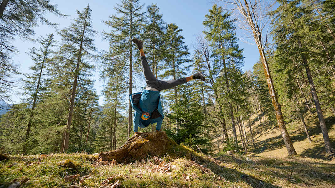 Person in Outdoorbekleidung von Maier Sports macht einen Handstand auf einem Baumstumpf, umgeben von einem sonnigen Wald.