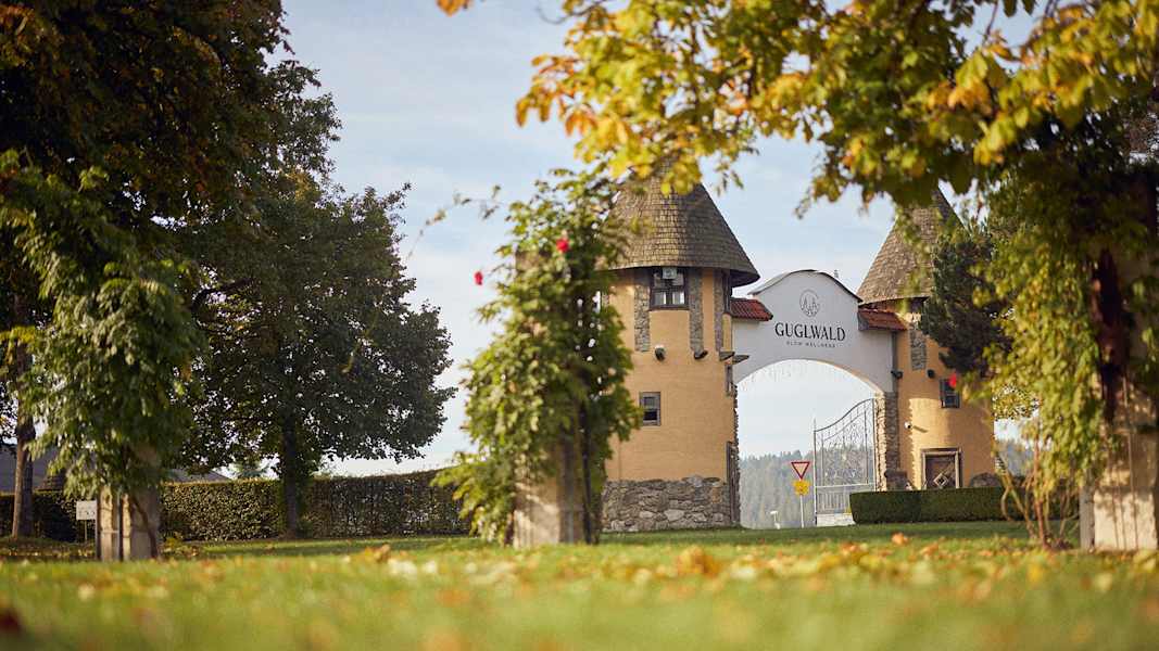 Blick auf das herbstlich umrahmte Eingangstor des Hotel Guglwald mit Bäumen und buntem Laub im Vordergrund.