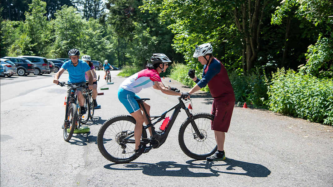 Mountainbike-Guide Georg Fröhlich zeigt einer Fahrerin beim Techniktraining im Freien die richtige Position auf dem Bike, weitere Teilnehmer:innen warten im Hintergrund.