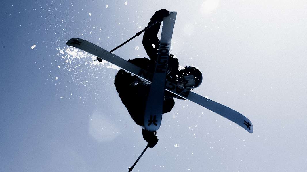 Freestyle-Skifahrer in der Luft bei einem Trick, mit Sicherheitsausrüstung und Ski unter blauem Himmel.