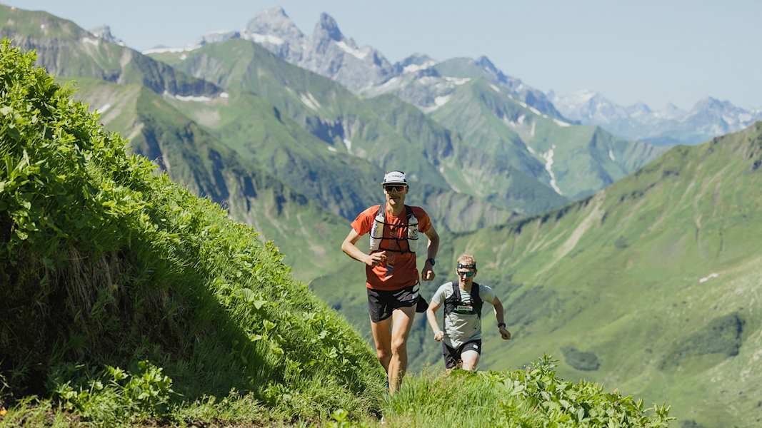 Zwei Trailrunner auf einem Bergpfad in Richtung Himmeleck, umgeben von grünen Hängen und den Gipfeln der Allgäuer Alpen.