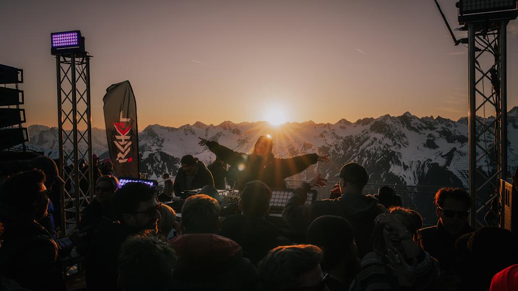 Eine Frau, die DJ ist, streckt ihre Arme aus und genießt die Musik sowie die Stimmung in Ischgl. Vor ihr steht eine tanzende Menge. Hinter ihr sieht man die Sonne hinter den Bergen untergehen.