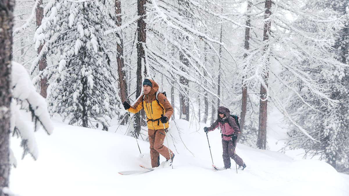 2 Personen gehen mit Ski und Ausrüstung durch den verschneiten Wald im Winter.