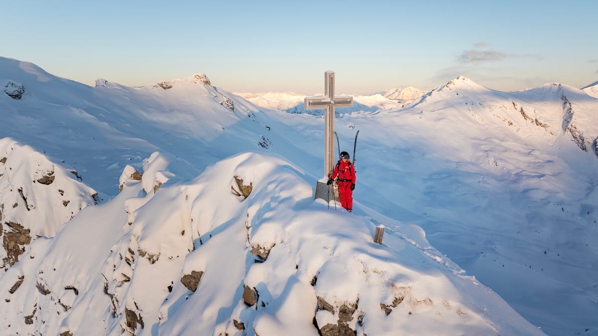 Ein Mann mit Skikleidung von Helly Hansen auf einen Schneebedeckten Berggipfel neben einen großen Kreuz.