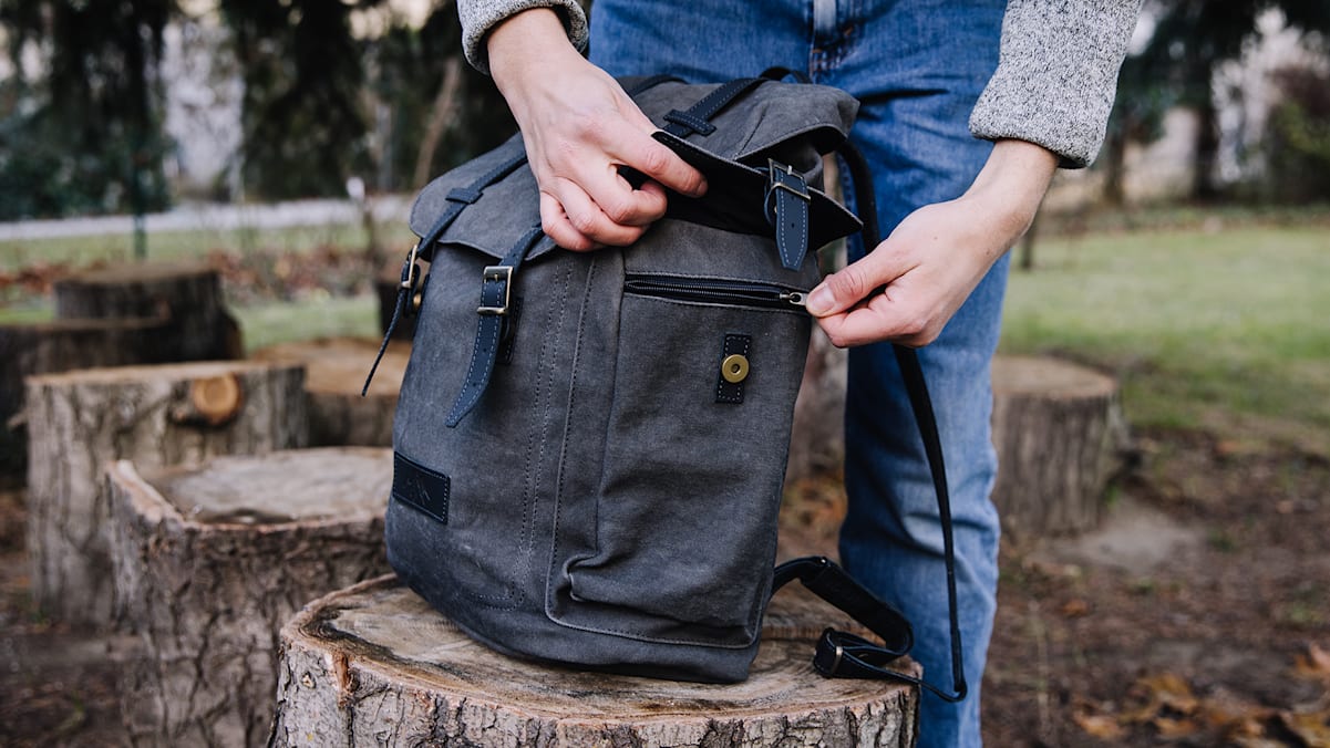 Ausgestattet mit Seitentaschen, Rückenpolsterung und viel Stauraum steht der graue Rucksack auf einem Holzstück im Freien.