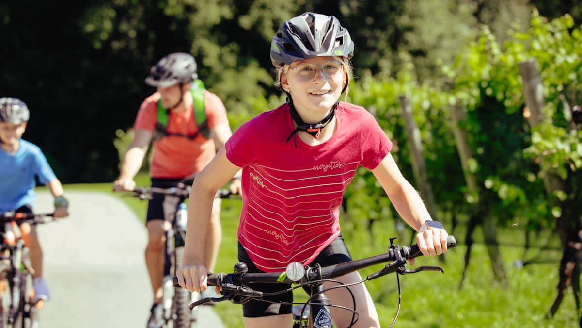 Ein junges Mädchen mit glücklichem Gesicht radelt an Weinbergen vorbei. Im Hintergrund ihr Vater und ihr Bruder.