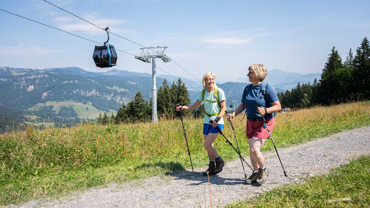 Zwei Wanderinnen mit Stöcken gehen auf einem Wanderweg unter einer Bergbahn.
