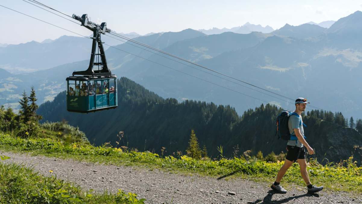 Ein Wanderer geht über eine Forststraße am Walmendinger Horn, im Hintergrund eine Gondel und Berge.