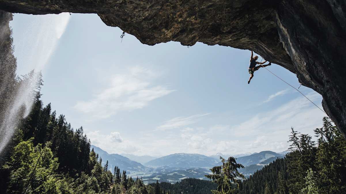 Im Vordergrund: ein Kletterer im Überhang und ein Wasserfall. Im Hintergrund: eine Berglandschaft.