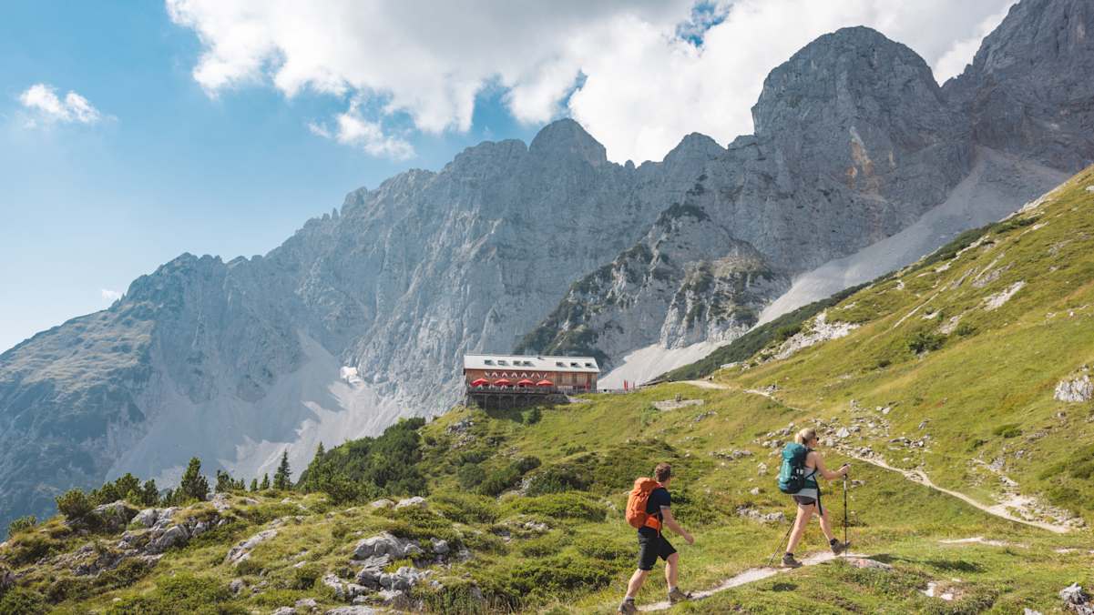 Zwei Wanderer nähern sich einer Alm, im Hintergrund steht eine Bergkette.