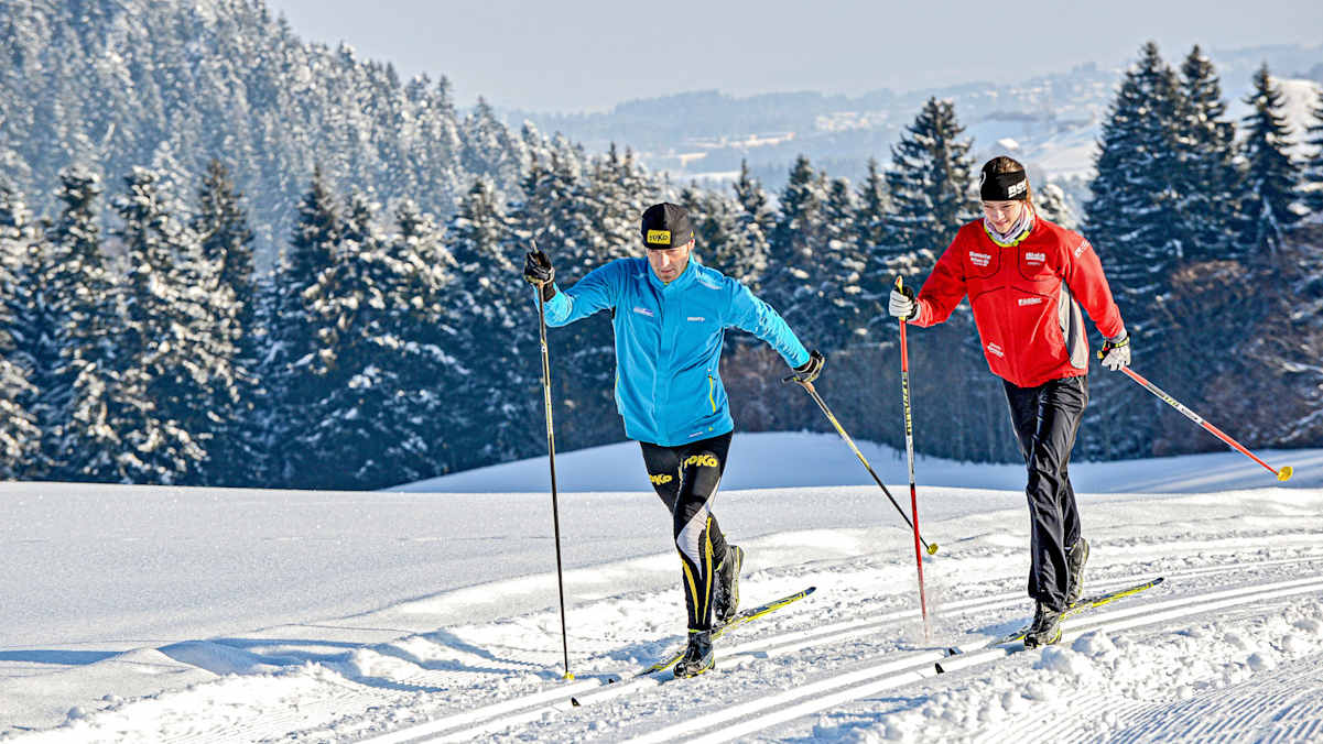 Zwei klassische Langläufer laufen nebeneinander durch die tief verschneite Winterlandschaft von Scheidegg im Allgäu.