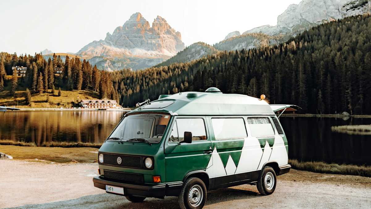 Ein weiß-grüner Campervan steht vor einem See, im Hintergrund eine angeleuchtete Bergkulisse.