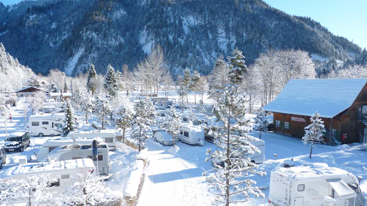 Blick auf den tief verschneiten Campingpark Oberammergau im Allgäu: Im Vordergrund verschneite Wohnwägen und Bäume, im Hintergrund eine bewaldete Bergkulisse.