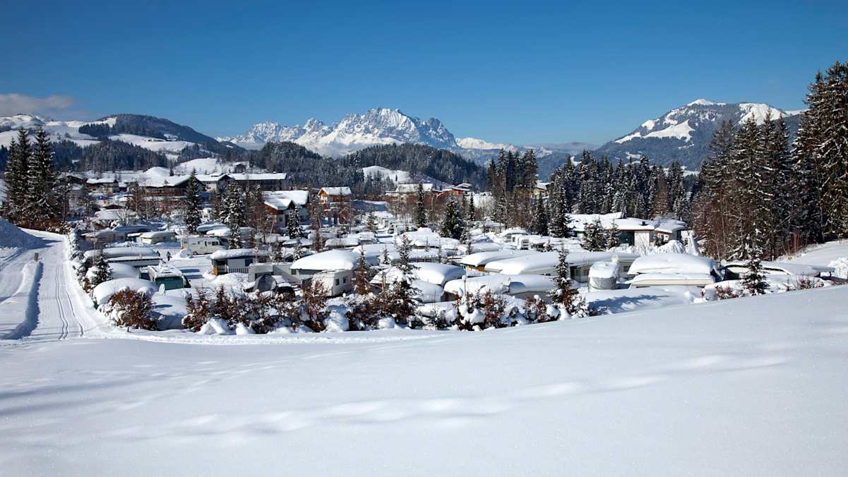 Blick in die tief verschneite Landschaft rund um das Tirol Camp in Fieberbrunn. Im Vordergrund die Wohnwägen, im Hintergrund eine verschneite Bergkulisse.