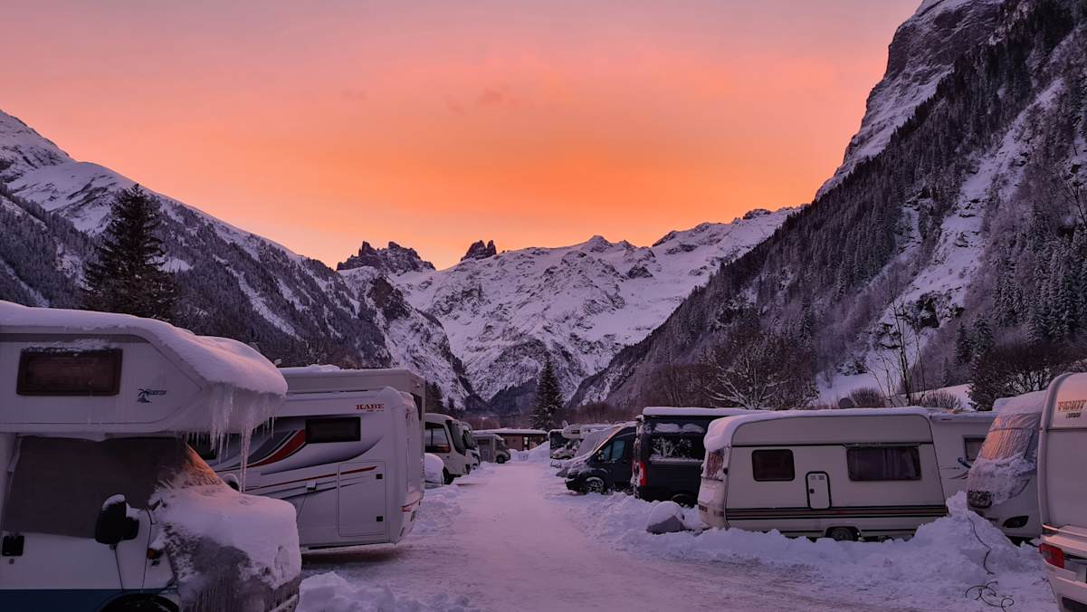 Sonnenuntergang im Alpenresort Eienwäldi Engelberg: Eingeschneite Wohnwagen und Camper stehen vor einer schneebedeckten Bergkulisse.