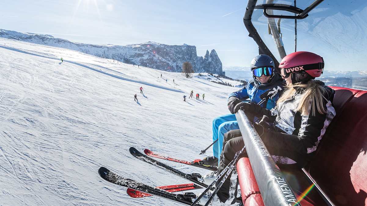 Ein Mann und eine Frau sitzen in einem Sessellift der Dolomitenregion Seiser Alm. Dahinter ist eine Skipiste und das Bergpanorama zu sehen.