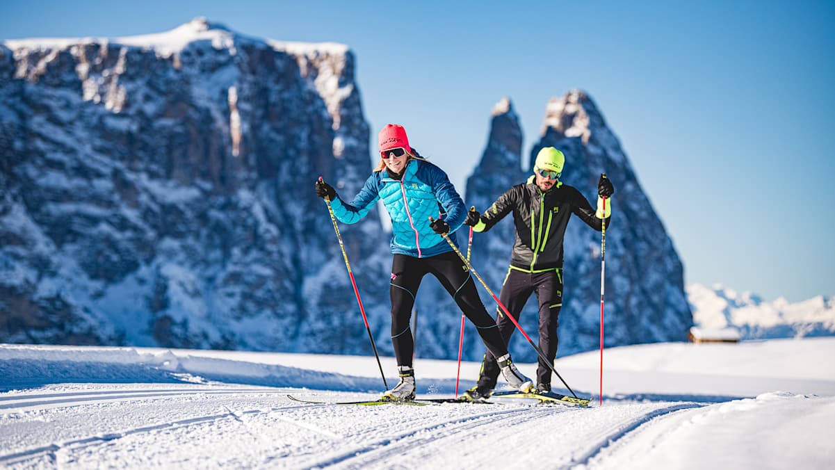 Ein eine Frau und ein Mann freuen sich über das Langlaufen auf einer der präparierten Loipen der Dolomitenregion Seiser Alm. Im Hintergrund sind markante Bergspitzen zu sehen.