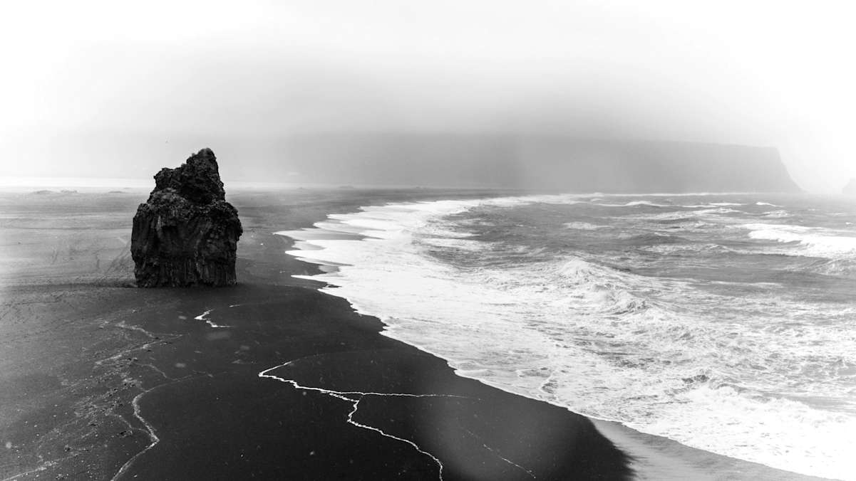 Einsamer Felsturm am Reynisfjara, dem Schwarzen Strand an der Südküste mit aufgewühltem Meer und nebligem Himmel