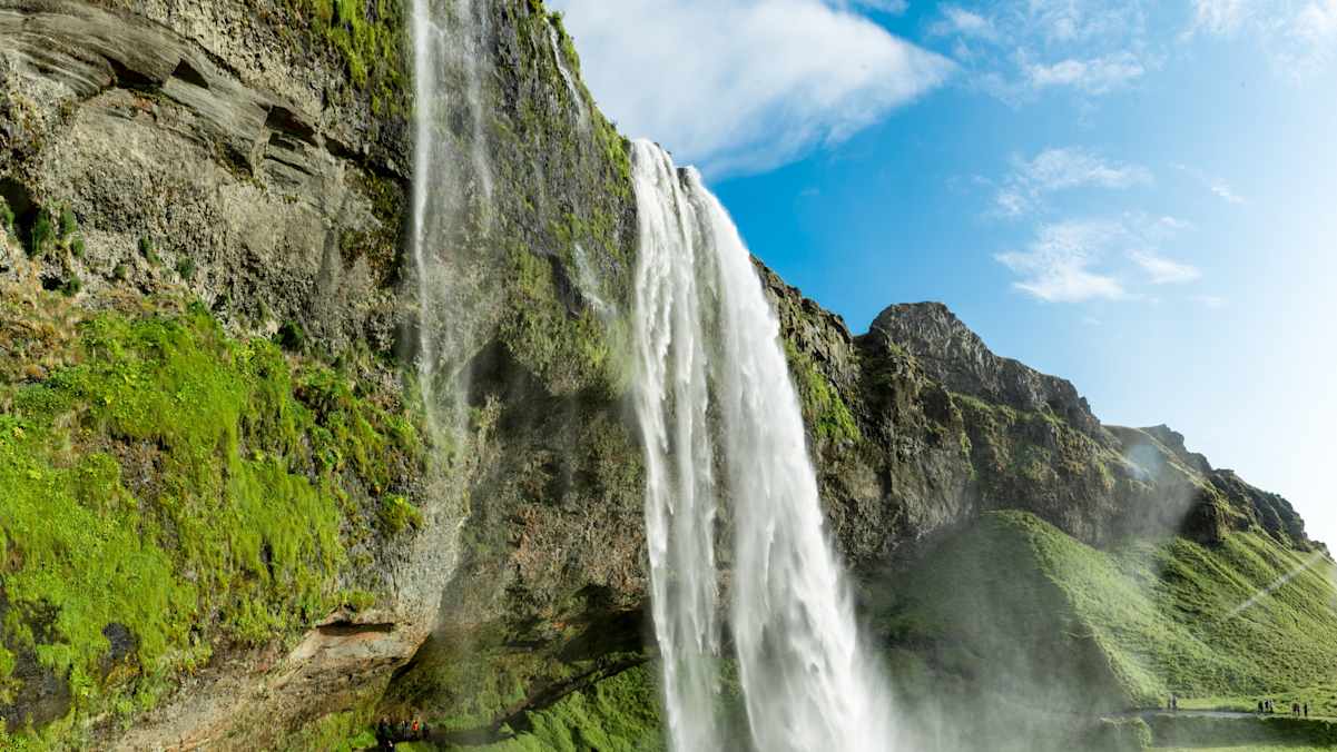 Der Seljalandsfoss an bewachsener Felsklippe vor blauem Himmel