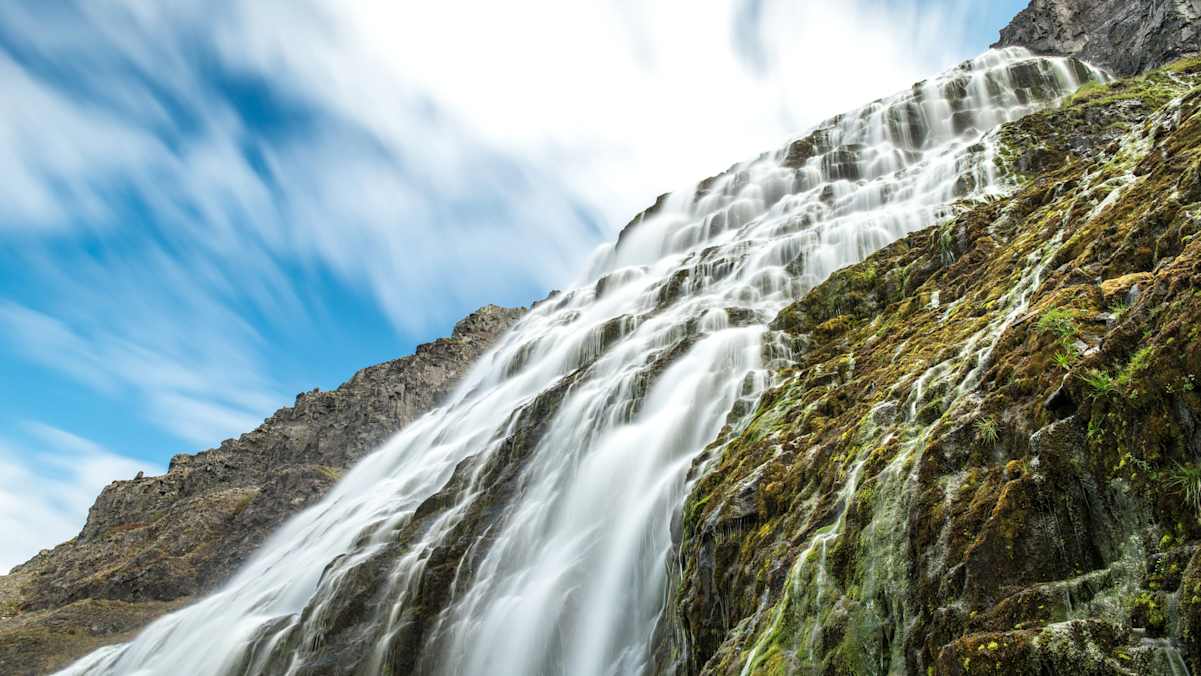 Wasserfall über bemoostem Fels vor leicht bewölktem Himmel
