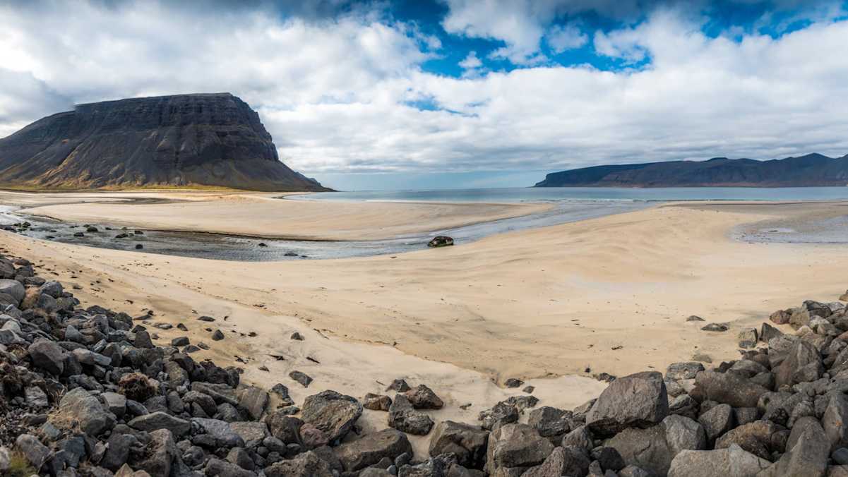 Heller Sandstrand mit dunklen Bergen und Meer im Hintergrund, im Vordergrund dunkles Gestein.