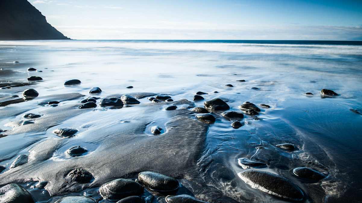 Dunkler Sand mit nassen Steinen bei Ebbe und Meer im Hintergrund