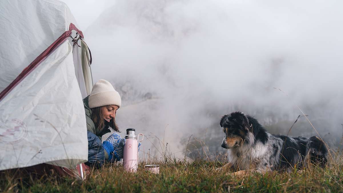 Frau im Zelt bei einer Bergwanderung mit Hund, umgeben von Nebel und Bergen, neben einer rosa Hydro Flask Hot Flask & Cup für heiße Getränke.