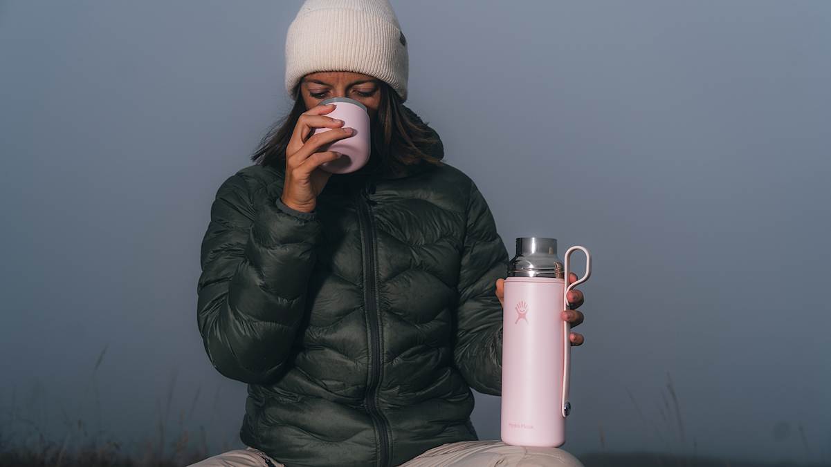 Frau in Winterkleidung genießt ein heißes Getränk aus der rosa Hydro Flask Hot Flask & Cup während einer Pause in nebliger Natur.