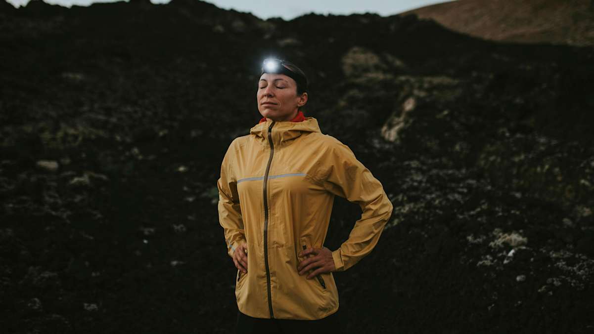 Eine Frau steht im Dunkeln mit einer Stirnlampe vor schwarzem Gestein.