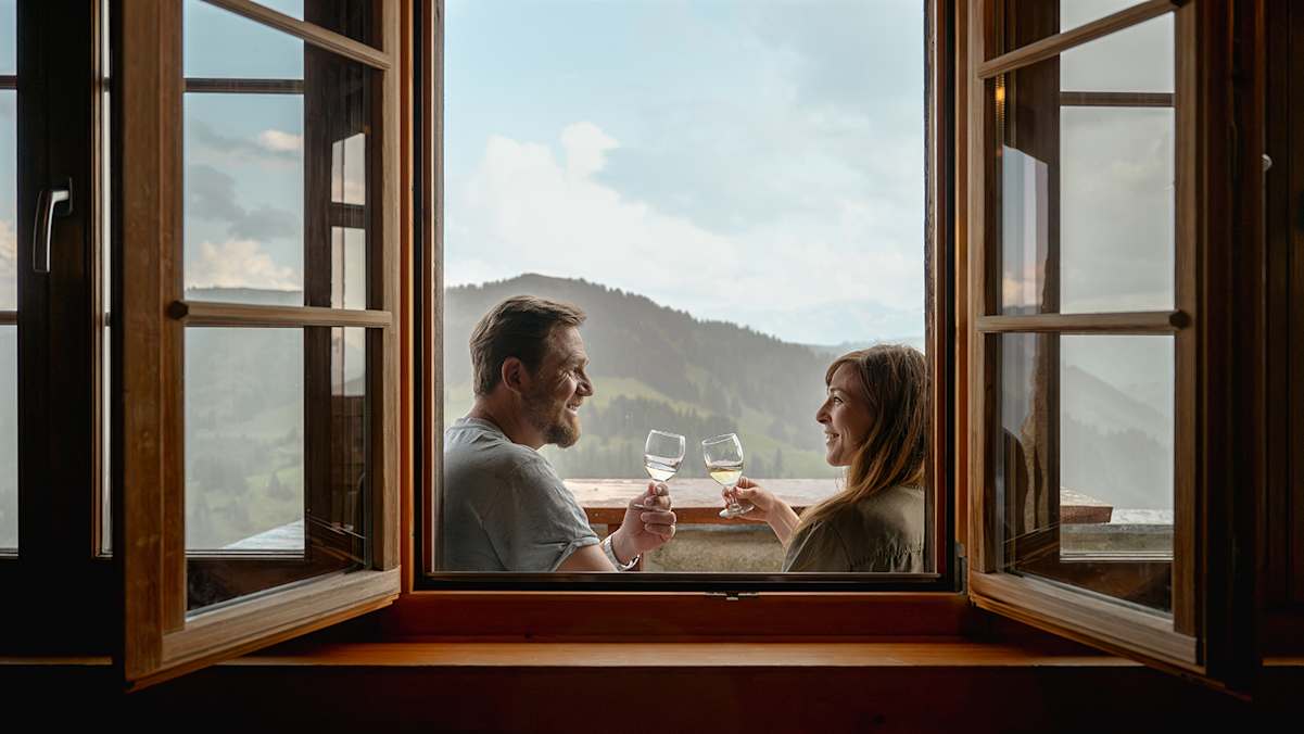 Ein Paar genießt ein Glas Wein mit Blick auf die Berglandschaft in Gstaad durch ein offenes Fenster.