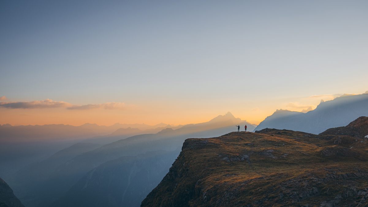 Kontrastreich: Layer für Layer präsentieren sich die Gipfel der Glarner Alpen im goldenen Licht der aufgehenden Sonne. Vor dem Horizont heben sich die zwei Personen am Berggipfel kontrastreich ab.