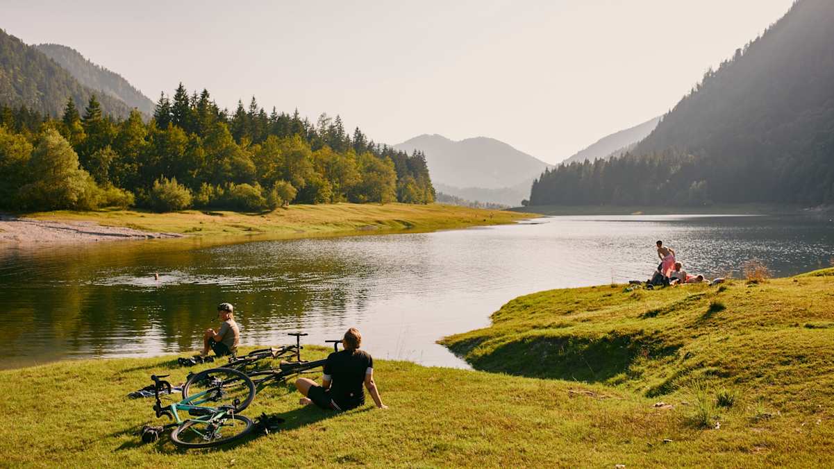 Drei Personen sitzen am grasigen Seeufer des Lödensee in Bayern bei goldenem Licht und schönem Wetter