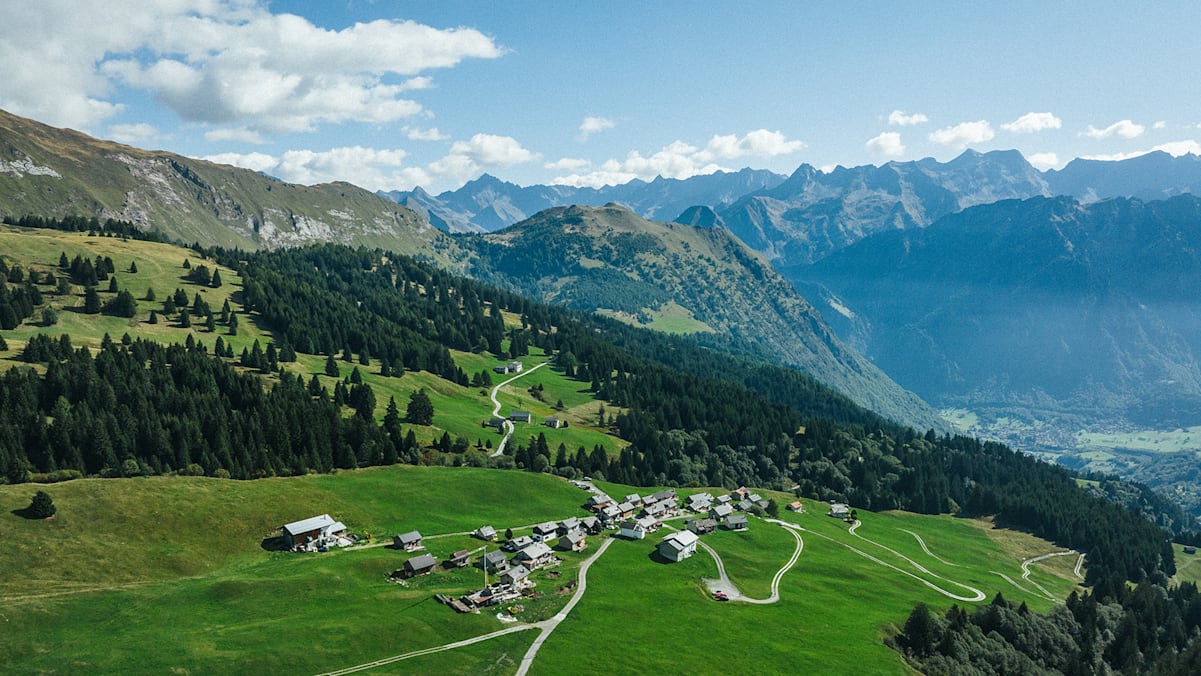Ein Bild von einer Ortschaft in den Schweizer Alpen und rundherum viele Wälder, Wiesen und Berge.