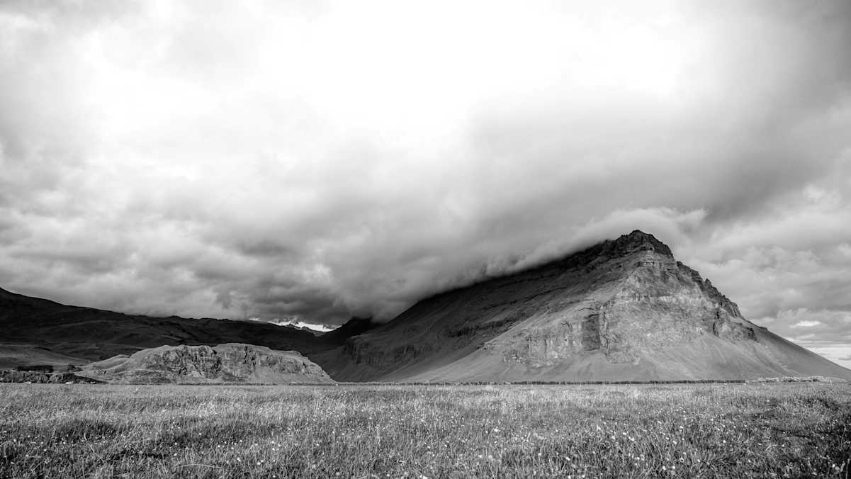 Eine Wiese vor karger Berglandschhaft mit dunklen Wolken, welche sich über den Bergen türmen.