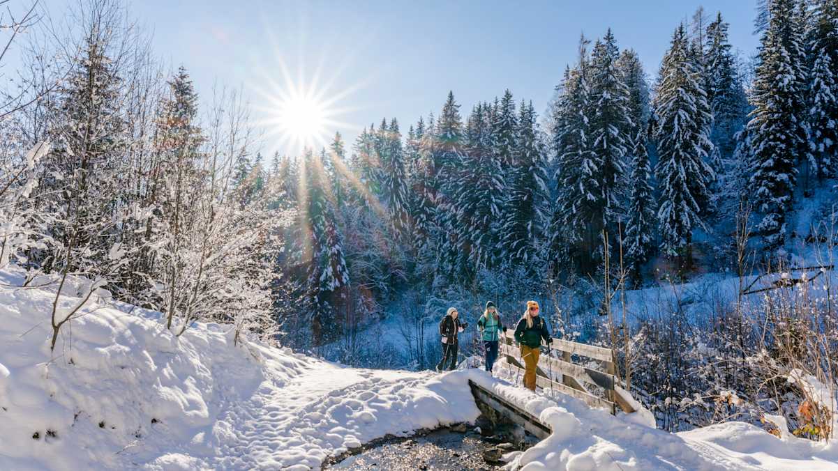 Drei Winterwanderer stapfen durch eine tief verschneite Schneelandschaft am Wilden Kaiser. Sie überqueren gerade eine eingeschneite Brücke über einen kleinen Bach.