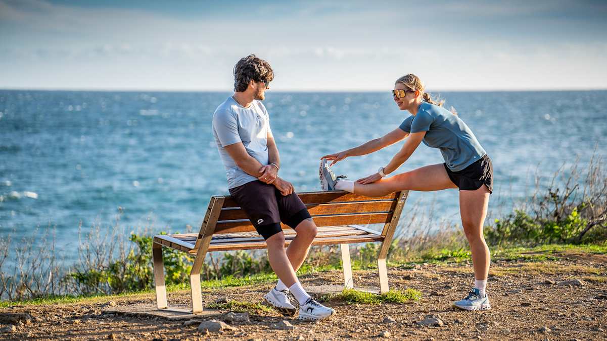 Eine Frau und ein Mann dehnen sich vor einer Bank. Sie tragen dabei Trailrunning-Outfits von skinfit. Hinter ihnen ist das Meer.