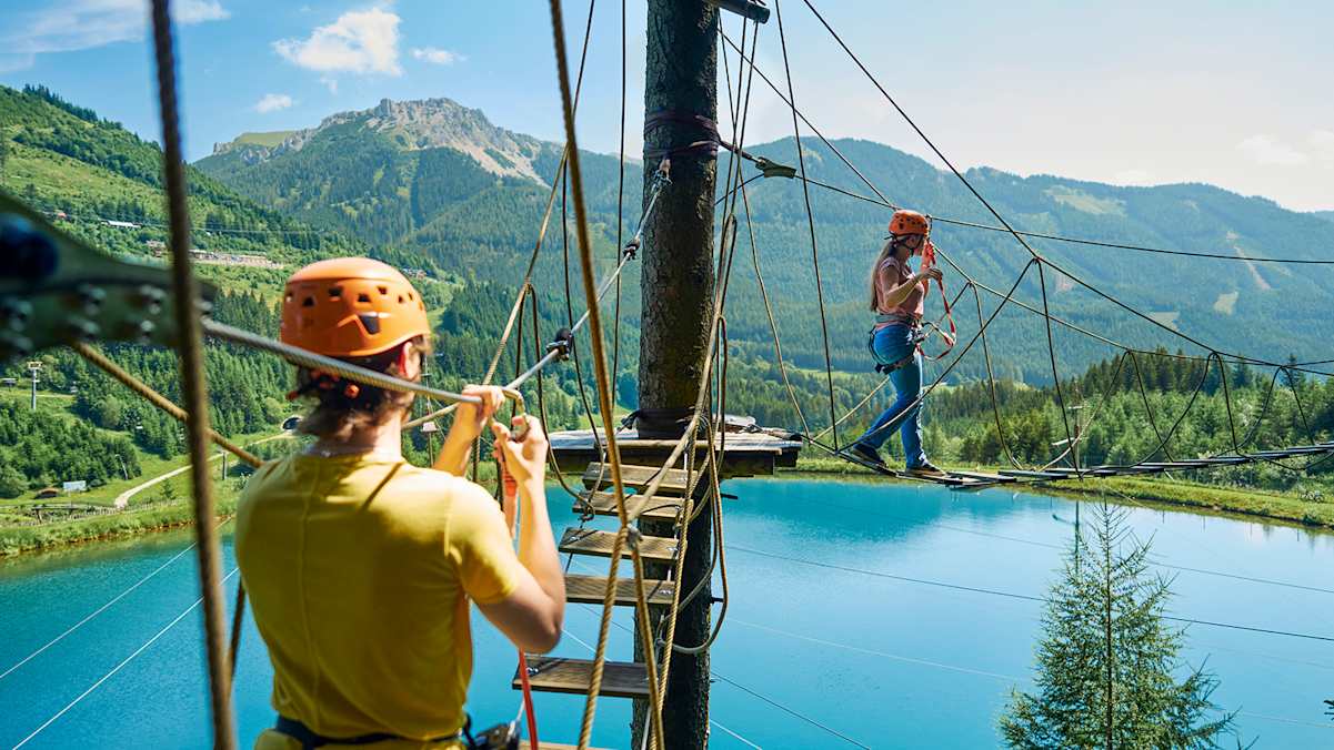 Zwei Personen im Hochseilgarten Alpfox überqueren eine Seilbrücke mit Blick auf den Grüblsee und die umliegenden Berge.
