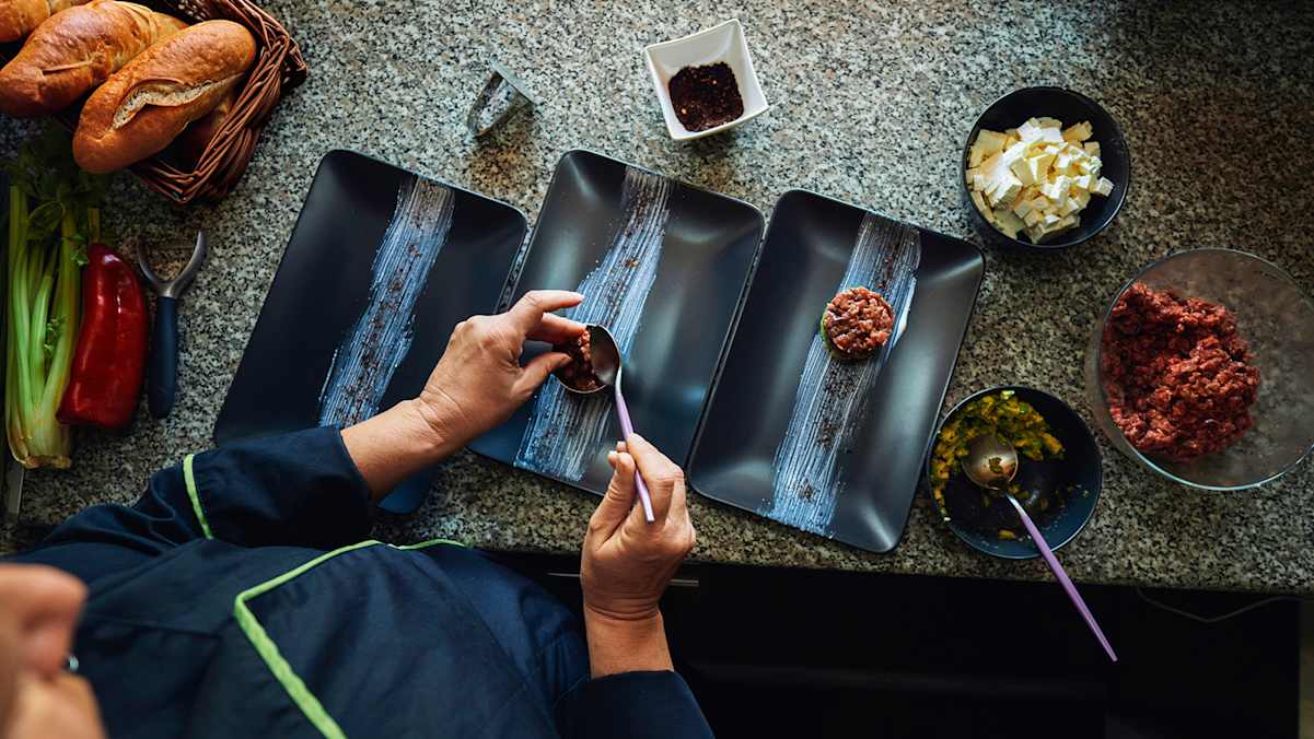 Eine Köchin richtet in der Seehütte am Grüblsee frisches Beef-Tatar auf schwarzen Tellern an, umgeben von Zutaten.
