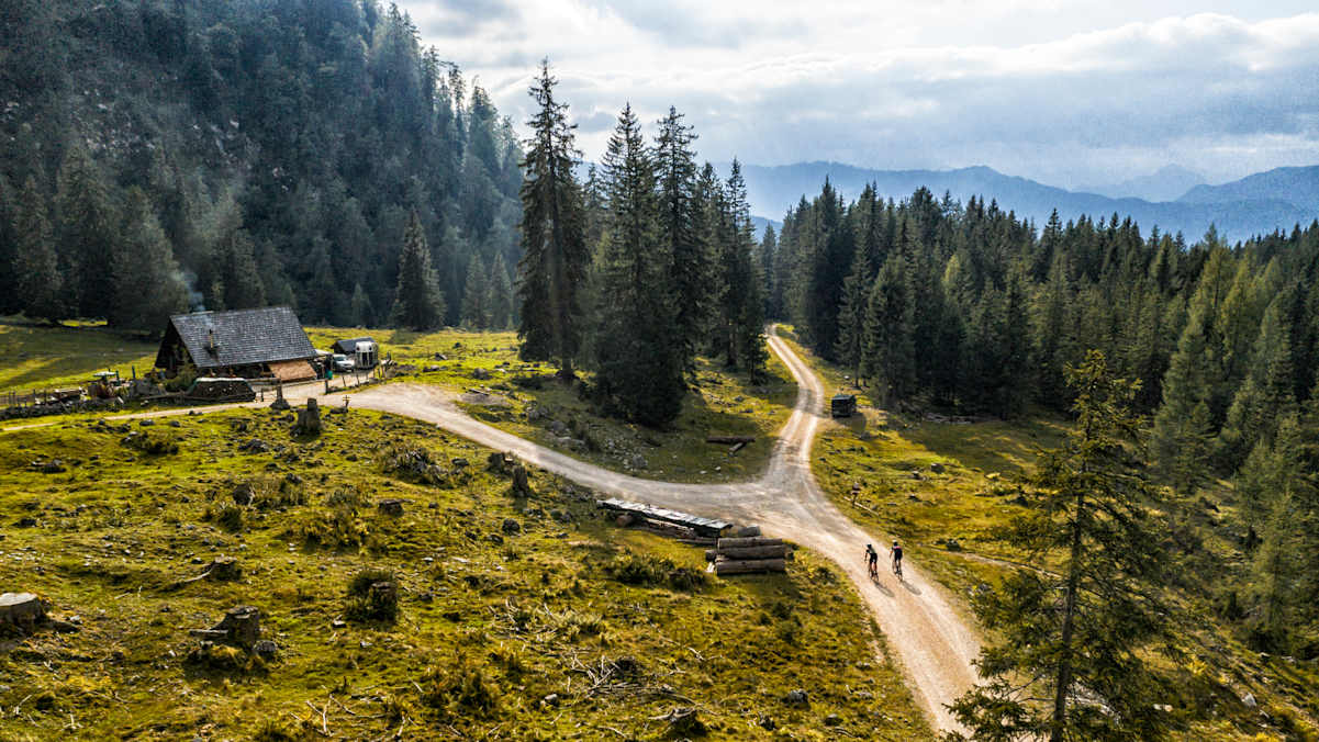 Zwei Gravelbiker fahren über eine Schotterstraße, im Hintergrund eine Almhütte.