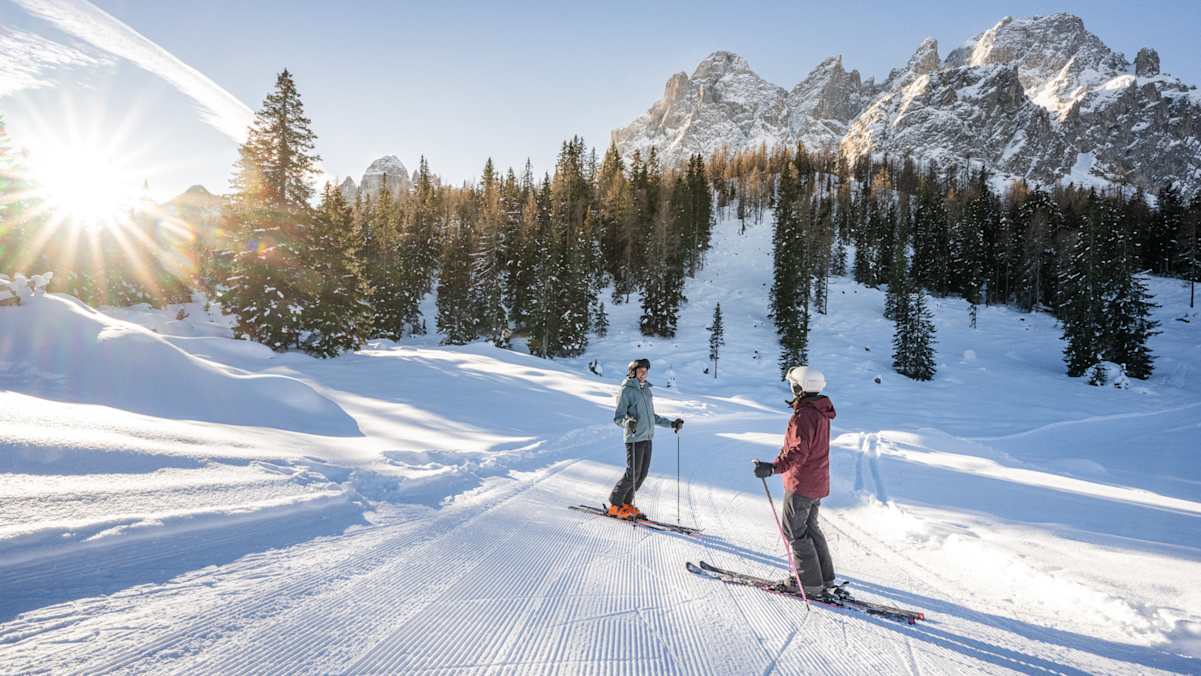 Zwei Skifahrer legen auf der Gran Giro delle Cime eine kurze Pause ein und genießen die tief verschneite Landschaft im Sonnenschein.