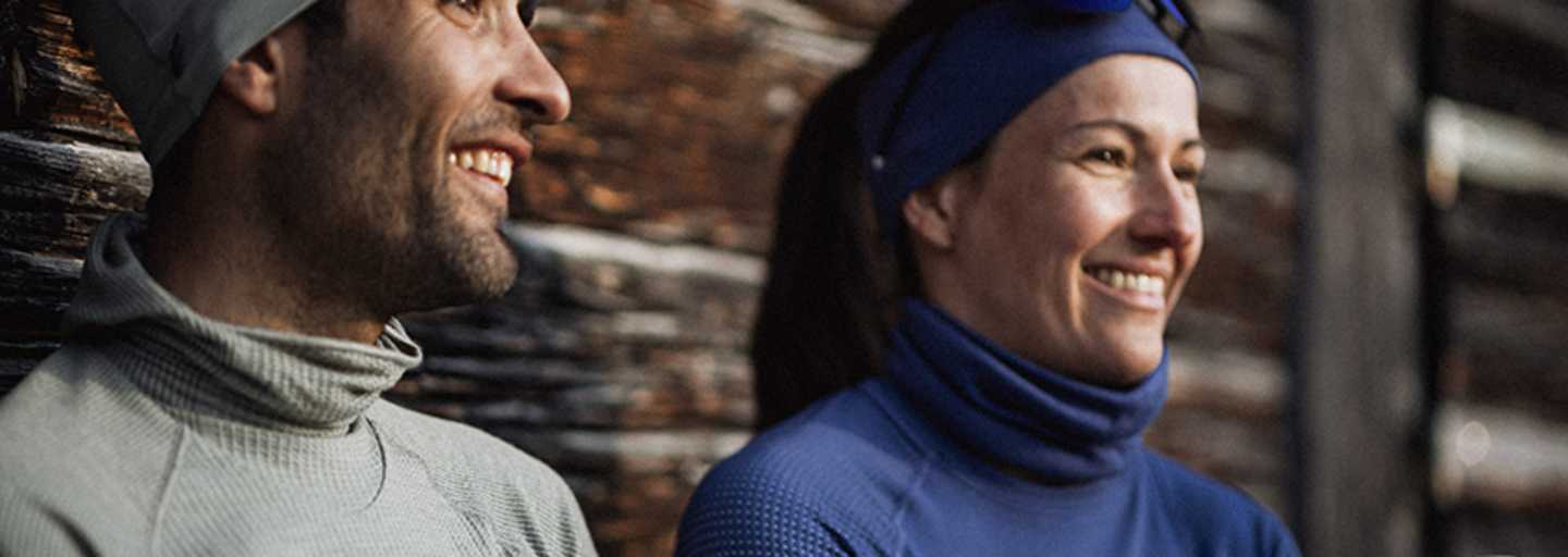 Zwei Personen in funktionaler Winterbekleidung von Odlo, genießen eine Pause im Freien vor einer rustikalen Holzhütte.